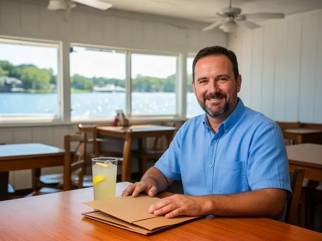 Smiling restaurant owner in a blue shirt with his hands on a closed folder at a table in an empty lakeside dining room